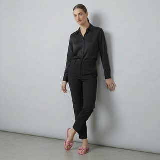 A female model leaning against a white wall wearing a black long-sleeve silk button-down shirt, black tailored trousers, and bright pink studded bow jelly sandals.