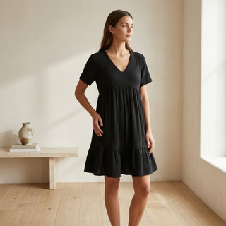 Woman wearing a black dress standing in a bright room with wooden floor and white walls.