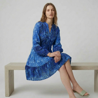 Woman wearing a blue patterned dress sitting on a wooden bench against a plain background
