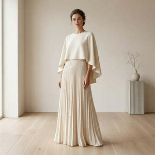Ultra-realistic lifestyle shot of a woman in a cream pleated maxi dress with a matching minimalist cape overlay in a bright, modern studio.