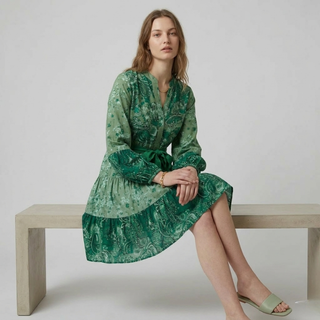 Woman wearing a green patterned dress sitting on a bench against a plain background