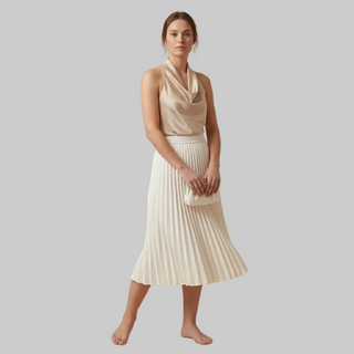 Woman wearing a beige sleeveless top and white pleated skirt on a beige background