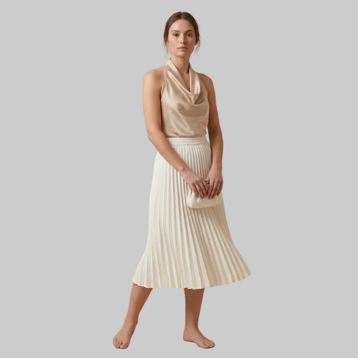 Woman wearing a beige sleeveless top and white pleated skirt on a beige background